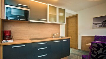A modern kitchen with dark cabinets and a wooden workspace. In the background, a table and a chair in purple can be seen.