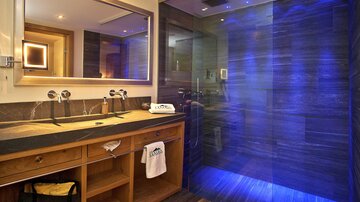 A modern bathroom with an elegant wooden vanity and a large mirror. The shower cabin has a glass wall with blue lights.