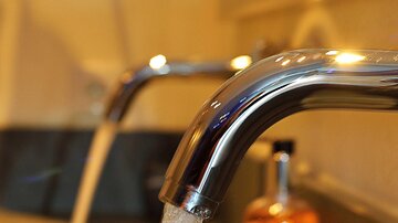 Two faucets, from which fresh water flows, are positioned above a modern sink. In the background, there is a bottle with an orange-colored liquid.