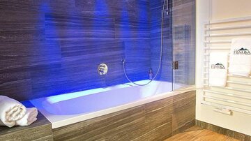 A modern bathroom with a freestanding bathtub and a rain shower. The walls are decorated with dark tiles and blue LED lights.
