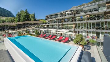 Ein modernes Hotel mit einem großen Außenpool und roten Liegen. Im Hintergrund sind grüne Berge und eine gemütliche Terrasse zu sehen.