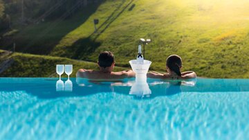 Ein Paar entspannt in einem Infinity-Pool mit Blick auf die grünen Wiesen. Neben ihnen stehen zwei Gläser und ein Flasche, die für einen besonderen Moment bereitsteht.