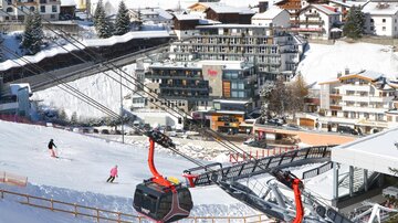 Eine malerische Ski-Landschaft mit schneebedeckten Pisten und einer modernen Gondelbahn. Im Hintergrund sind Berghotels und Fichten zu sehen.