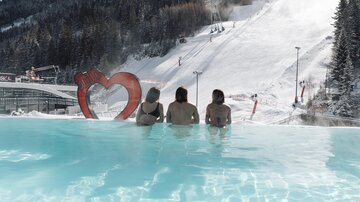 Ein warmer Pool mit Blick auf eine schneebedeckte Piste. Drei Personen genießen die Aussicht in einer entspannten Atmosphäre.