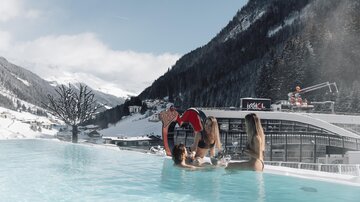 Ein beheizter Außenpool in den Bergen, umgeben von schneebedeckten Gipfeln. Drei Frauen genießen das Wasser und die Aussicht in einer entspannten Atmosphäre.
