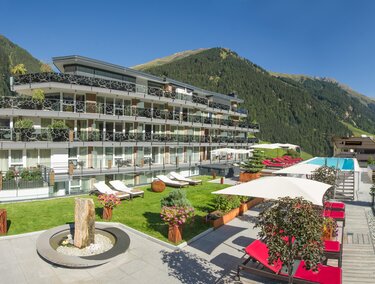 Ein modernes Hotelgebäude mit Balkonen und einem Pool in einer schönen Berglandschaft. Der Außenbereich ist mit Liegen und Pflanzen gestaltet.
