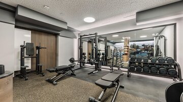 Ein moderner Fitnessraum mit verschiedenen Trainingsgeräten und Gewichten. Die Wände sind hell und Spiegel reflektieren den Raum.