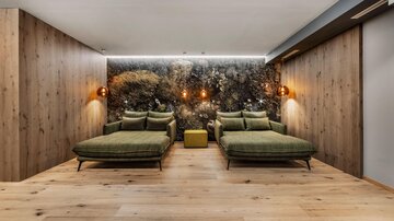 Ein moderner Entspannungsraum mit zwei grünen Liegen und warmen Holzdetails. Die Wand ist mit einer naturnahen Bepflanzung dekoriert, was eine beruhigende Atmosphäre schafft.