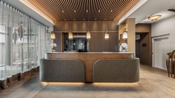 Eine elegante Rezeption mit stilvollem Holzdesign und modernen Lampen. Im Hintergrund sind große Fenster mit Vorhängen zu sehen, die für eine helle Atmosphäre sorgen.