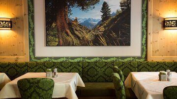Ein gemütliches Restaurant mit grünen, gemusterten Sitzbänken und Tischen. An der Wand hängt ein großes Bild einer Berglandschaft.