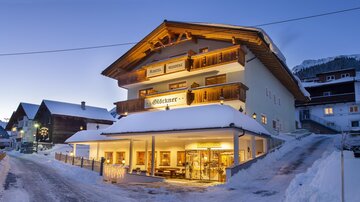 Ein charmantes Hotel im Schnee mit einem einladenden Eingang. Die Berge sind im Hintergrund sichtbar und der Himmel ist klar und blau.
