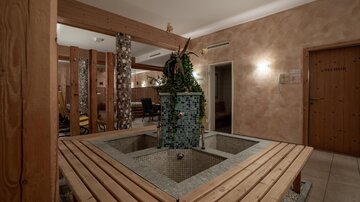 Ein gemütlicher Wellnessbereich mit einem zentralen Brunnen und Holzbank. Die Wände sind in warmen Erdtönen gehalten, und Pflanzen sorgen für eine entspannte Atmosphäre.
