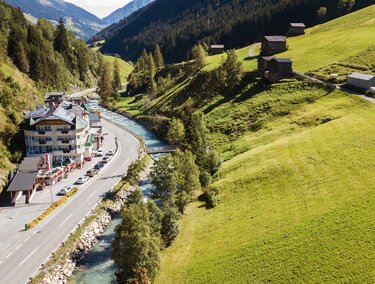Eine malerische Landschaft mit grünen Wiesen und einem kleinen Fluss. Im Hintergrund sind Berge und einige Holzgebäude zu sehen.
