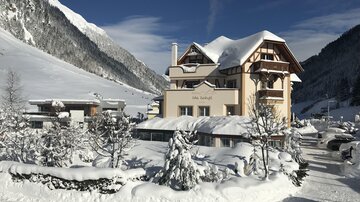 A beautiful building in the snow, surrounded by snow-covered trees. In the background, impressive mountains and a clear blue sky can be seen.