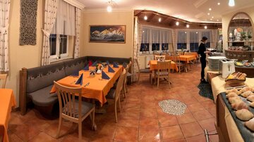 A cozy restaurant with orange tables and chairs. In the background, there are windows with a view and a buffet with fresh bread.