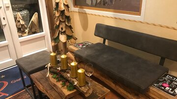 A cozy room with a wooden bench and table. On the table are candles and a glass, while a decorative tree design is visible in the background.