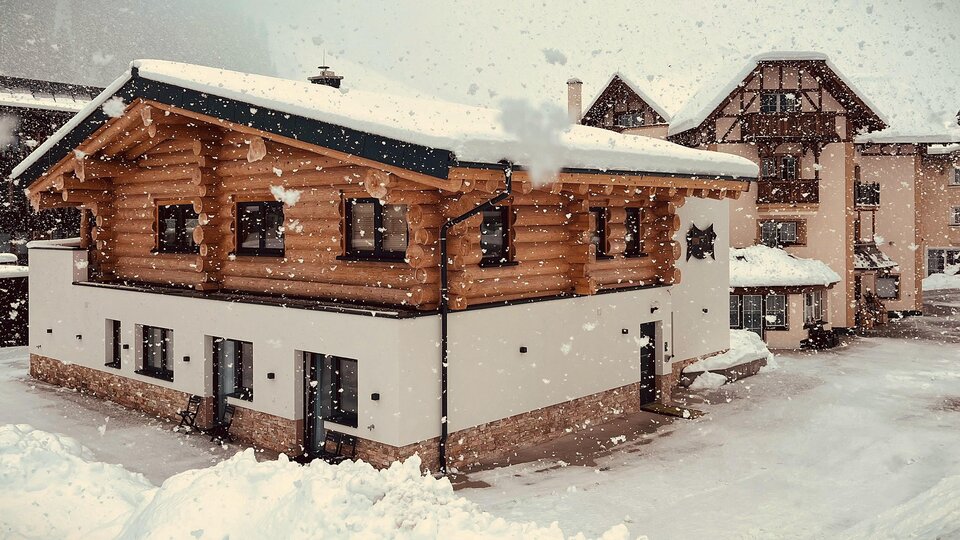 A charming wooden house in the snow, surrounded by a wintry landscape. Gentle snowflakes fall on the buildings in the background.