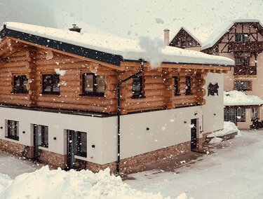 A charming wooden house in the snow, surrounded by a wintry landscape. Gentle snowflakes fall on the buildings in the background.
