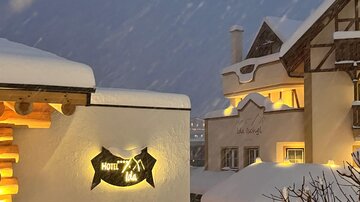 A snowy hotel with a glowing sign in the cold winter landscape. The buildings are designed in a traditional style and are surrounded by falling snow.