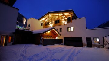 A cozy house in the snow, illuminated by warm light. The sky is dark blue, creating a relaxed atmosphere.