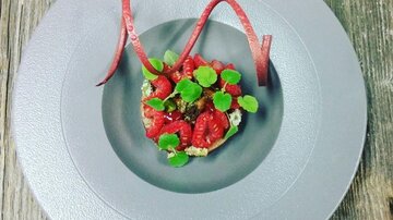 Ein kunstvoll angerichtetes Gericht auf einem grauen Teller. Es enthält frische Himbeeren, Gemüse und dekorative Garnituren.