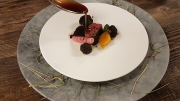 Ein elegantes Gericht auf einem weißen Teller, garniert mit buntem Gemüse und Saucen. Der Teller ist auf einem rustikalen Holzuntergrund präsentiert.