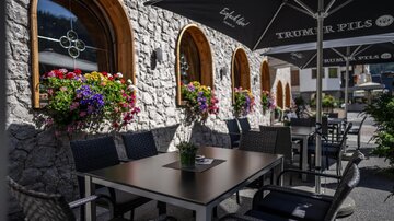 Ein gemütlicher Außenbereich mit einem Tisch und Stühlen vor einer steinernen Wand. Blumen in bunten Kästen schmücken die Fenster.