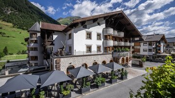 Ein charmantes Hotel in den Bergen, umgeben von grünen Wiesen und beeindruckenden Bergen. Die Terrasse mit Sonnenschirmen lädt zum Entspannen ein.