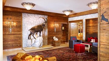 Eine einladende Lobby mit Holzwänden und einem großen Steinbock an der Wand. Stilvolle Möbel und ein Teppich verleihen der Umgebung eine gemütliche Atmosphäre.