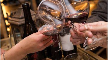 Mehrere Gläser mit Rotwein stoßen miteinander an. Im Hintergrund sind Weinflaschen und Korken sichtbar.