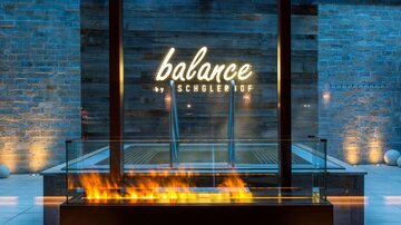 Ein eleganter Wellnessbereich mit einem gläsernen Wasserbecken und einer stilvollen Feuerschale. Der Schriftzug „balance“ ist prominent beleuchtet und sorgt für eine entspannende Atmosphäre.