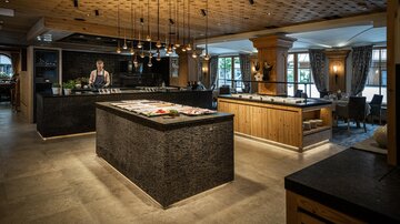 Ein modernes Restaurant mit Holz- und Steindekor. Im Vordergrund steht eine große Theke mit frischen Lebensmitteln und einem Koch.