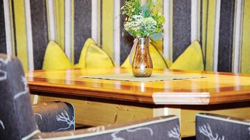 Ein gemütlicher Tisch mit einer blumigen Vase inmitten von bequemen Stühlen und Kissen. Die Wände sind mit gestreiftem Stoff in Gelb- und Grautönen dekoriert.