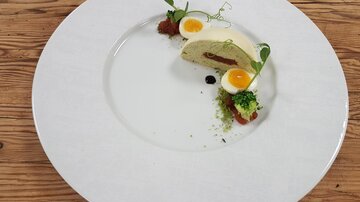 Ein eleganter Teller mit einer kreativen Speise, die ein Stück Terrine und zwei rohe Eier zeigt. Das Gericht ist mit frischen Kräutern und einer bunten Sauce garniert.