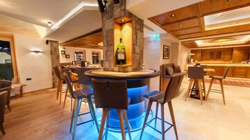 An elegant bar with stylish wooden chairs and a lit counter. In the background, shelves with drinks can be seen.