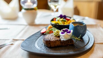 A beautifully arranged steak on a black plate, garnished with a flower and a sauce. Next to it is a bowl of colorful vegetables.