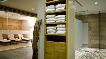 A modern wellness area with a wooden shelf displaying folded towels. In the background, there are loungers and a shower visible.