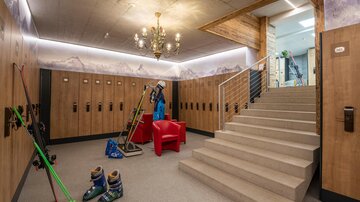 A modernly designed ski room with lockers and ski equipment. In the background, a staircase and a cozy chair can be seen.