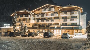 A hotel in the snow at night, illuminated by warm light. Parking spaces are available in front of the building.