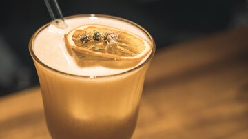 A refreshing cocktail with a slice of lemon and herbs. The drink is on a wooden surface and has an appealing, creamy look.