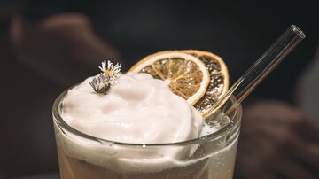 A delicious cocktail with a creamy foam crown and a slice of dried lemon. At the top, a small flower is decorated, and a straw completes the drink.