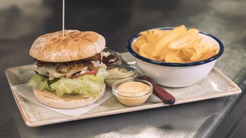 A juicy burger with lettuce, tomatoes, and onions on a plate. Next to it are crispy chips and a small bowl of dip.