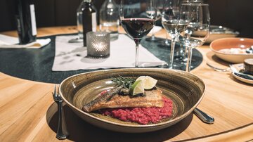An elegantly arranged dish featuring fried fish on a herb risotto with a slice of lime. In the background, wine glasses and other dishes can be seen.