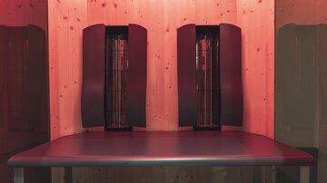 A wellness room with two infrared heaters and a smooth, dark lying surface. The walls are made of light wood and the room is softly illuminated in red.