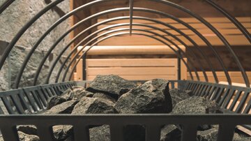 A sauna stove with stones in a metal frame. The background shows a wooden bench and a pleasant, relaxed atmosphere.