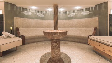 An elegant wellness area with warm stones and cozy loungers. In the center stands a stylish fountain, surrounded by soothing colors.