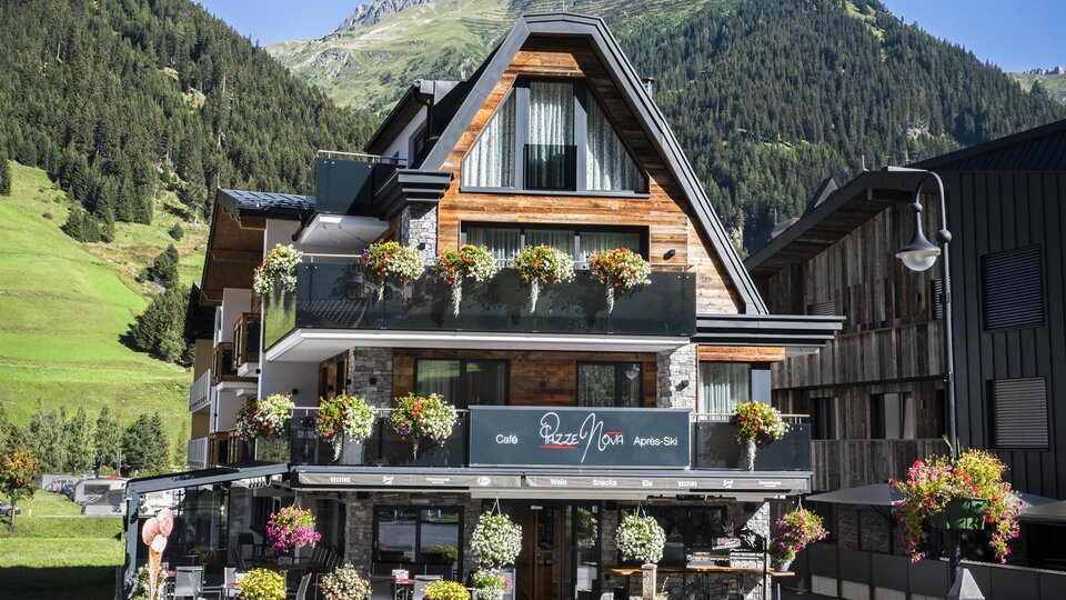 Ein modernes Chalet mit blühenden Pflanzen und einem großen Balkon. Im Hintergrund sind grüne Berge und ein klarer Himmel zu sehen.