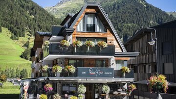 Ein modernes Chalet mit blühenden Pflanzen und einem großen Balkon. Im Hintergrund sind grüne Berge und ein klarer Himmel zu sehen.