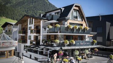 Ein modernes Hotelgebäude mit Balkonen und Blumendekoration. Im Vordergrund fahren Radfahrer vorbei, umgeben von einer schönen Berglandschaft.