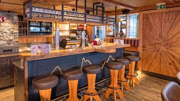 Eine gemütliche Bar mit Holztheke und modernen Hockern. Der Raum ist warm und einladend gestaltet.